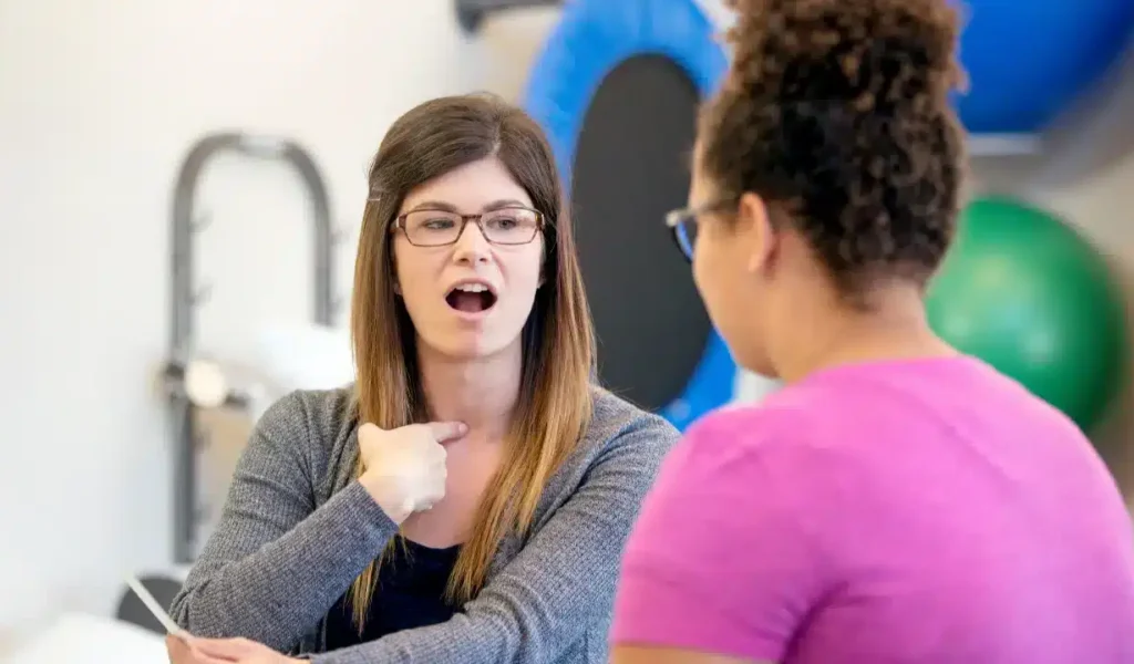 A woman in glasses and a pink shirt engages in conversation with another woman, discussing AI speech therapy.